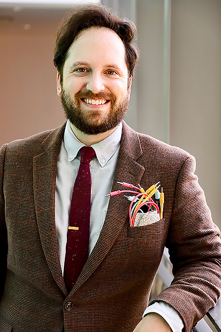 Anthony Marasco stands wearing a brown suit and red tie, elbow leaning on a partition. 