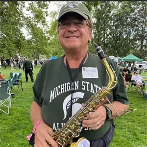 Augie Thoma holding his saxophone, wearing Michigan State University shirt and hat, standing in a field on campus with tailgaters in the background