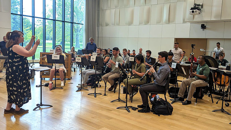 Conductor standing to the left of a wind ensemble, directing musicians while they perform. Setting, large rehearsal hall with windows letting in lots of light