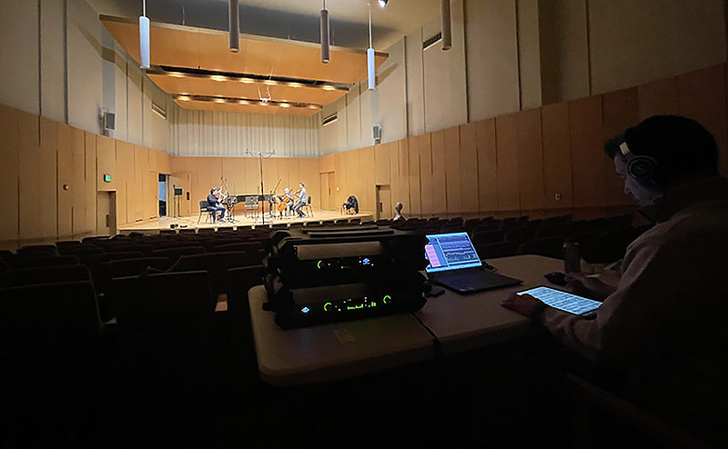 sound technician, seated, monitors recording equipment as he watches performers on stage play to an empty hall.