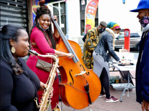 a saxophonist and bassist perform outdoors in a city while a man watches and people serve food in the background.