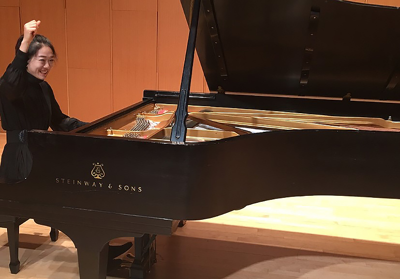 pianist, seated at piano in recital hall, smiles and raises right hand in the air at the end of a performance.