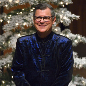 David Rayl, blue suit, smiles on stage with holiday tree in background