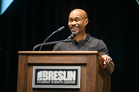 Man, smiling, speaks at wooden podium with a sign on it that says Breslin Student Events Center