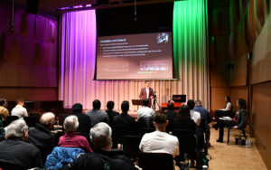 Audience gathered in Murray Hall, dimly lit venue with project slide image and colorful lighting, student is presenting a music business idea