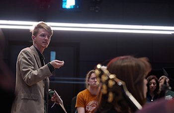 in a rehearsal hall full of students, one of them stands and gestures to another while speaking to the group