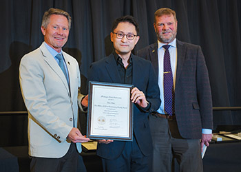 Three men pose for a photo, dressed in business suites, with the one on the left holding and presenting a plaque to the man in the center who also holds the plaque.