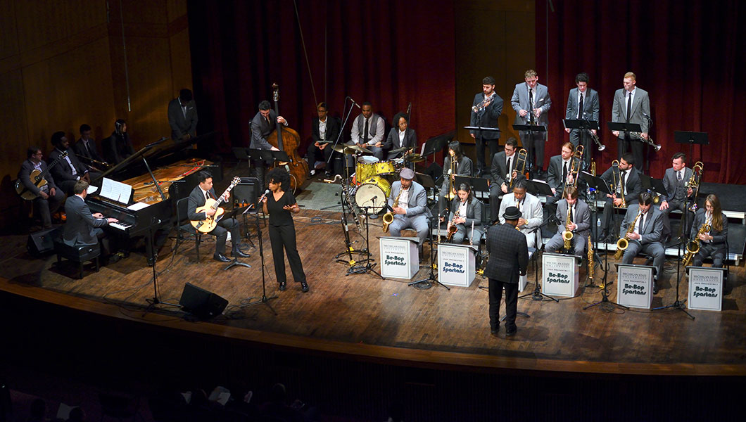 Full MSU Jazz Orchestra on stage of the Fairchild Theatre, MSU Auditorium. Dramatic stage lighting, formally dressed ensemble intently playing music. Directed by a conductor and a jazz vocalist sings to audience.