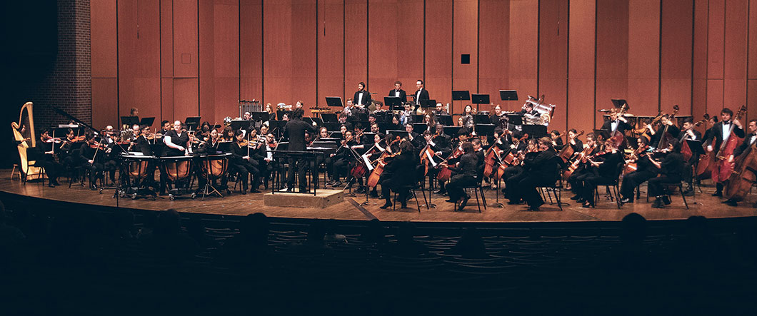 Symphony orchestra members fill Cobb Great Hall stage of Wharton center, dressed in formal attire, performing with conductor in foregrounc