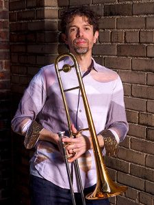 Photo portrait of jazz trombonist Ryan Keberle. Standing along a brick wall, casual attire and pleasant facial expression, looking at camera