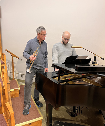 two men look at sheet music, one on left holding a trumpet and the one on right playing the piano.