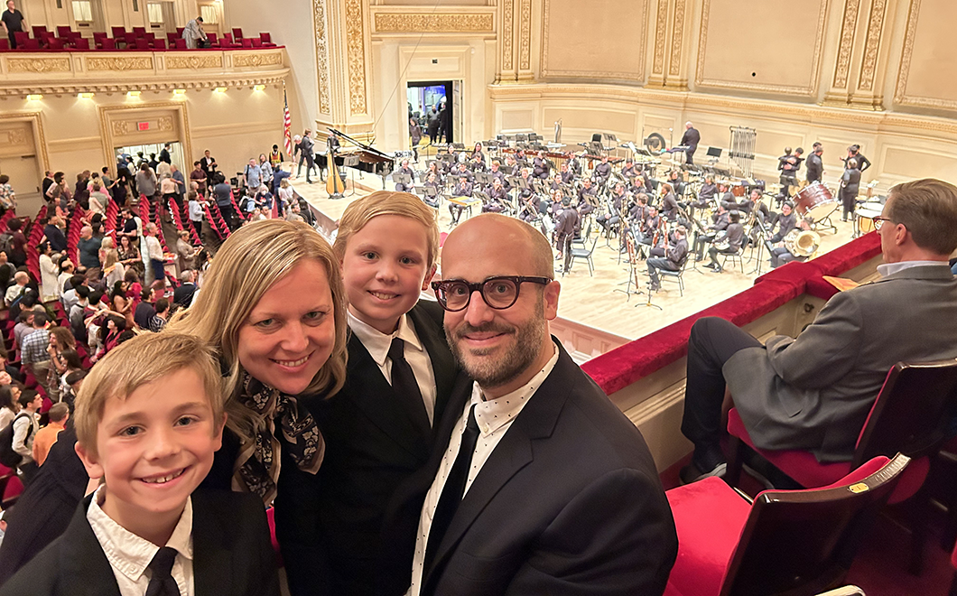 Young family of four poses and smiles as two sons, mother and father look at camera while seated in a balcony with an orchestra on stage in the background.