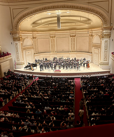 A long view of musicians on stage at Carnegie Hall, taken from up high in a balcony, the bright white/ivory stage accented by the red carpet of the theatre.