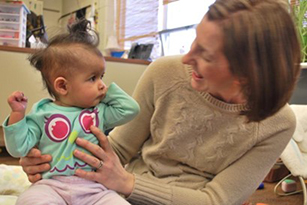 woman, right, smiles at a baby she holds up on her knee.