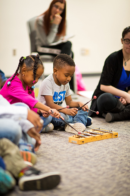 Two young children sit on floor playing a xylophone together, among a class of adults and children, while an adult in the background observes.