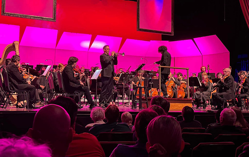 A view of an ensemble on stage from close seats on the left of the stage. On stage is a trumpeter and conductor with a large group of other musicians. The stage is lit brightly in pink hues.