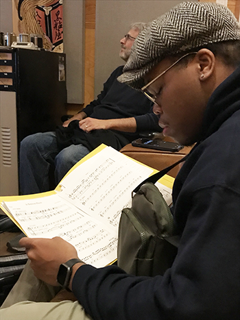 Close up of man, right, wearing a hat and glasses, looking closely at sheet music, right. 