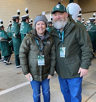 woman and man pose outside a stadium, dressed in warm clothing, with marching band members in the background