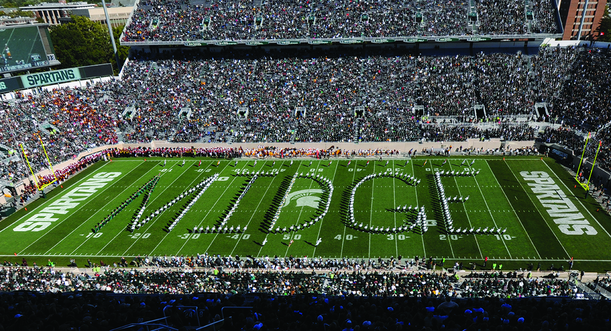 Football stadium, filled with people; on the green field, a marching band spells out the name Midge.