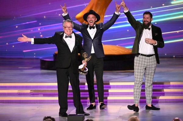 three men on stage, waving their arms and smiling, celebrating with the Emmy Award trophy they each hold.