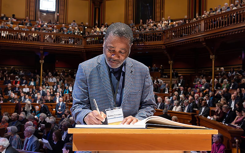 Close up of man writing in a large open book on a podium with a crowd of people in an old auditorium behind him