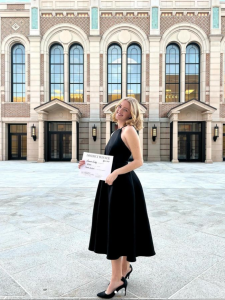 Woman in black dress stands outdoors in front of a building with large windows rounded at the top. She smiles and holds a certiicate.