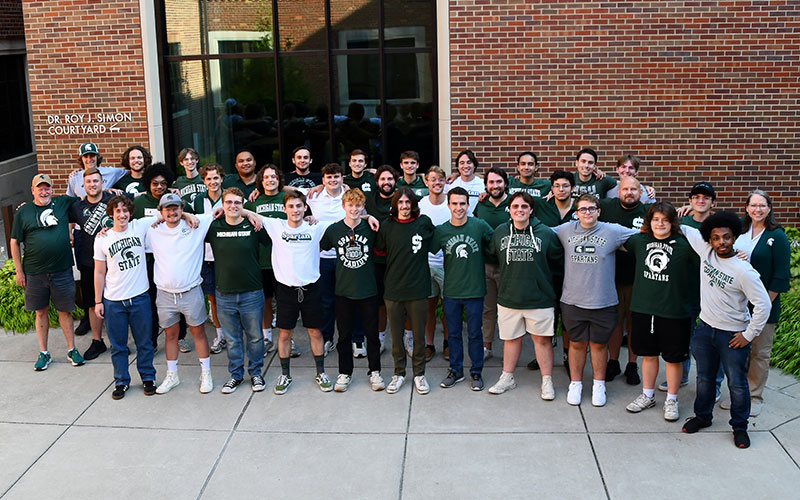 Gathering of 35 male singers for a group picture of the choral group Signing Spartans. Members looking at camera and smiling, arms joined over shoulders and wearing MSU Spartan attire