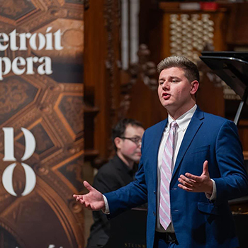 Young man in blue sport coat sings on stage with partial words on banner reading Detroit Opera.