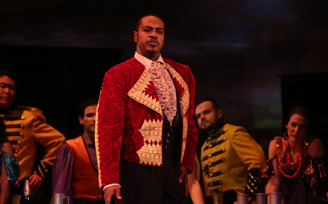 A man stands in red, regal costume on an opera stage during a performance.