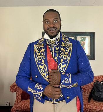 Man smiles at camera, standing in a dressing room while wearing an ornate, blue opera costume.