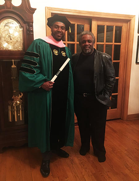 Two men smile as one wears college graduation cap and gown, holding a diploma.