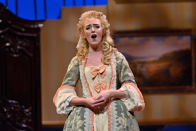 woman in ornate, pale green and peach dress sings an opera role on stage.