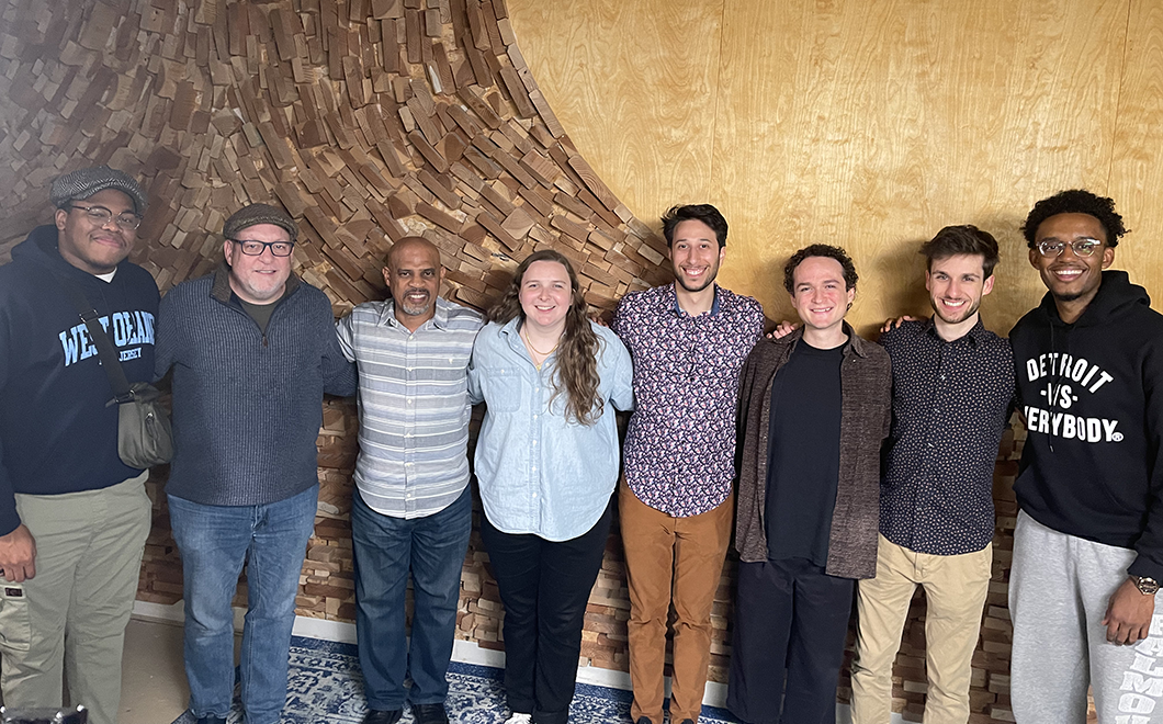 Eight people stand shoulder to shoulder, smiling, with a wooden wall design behind them.