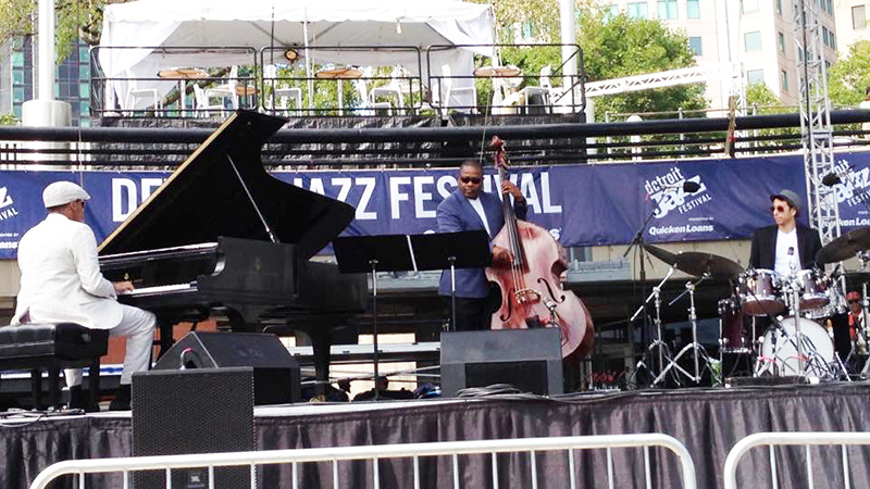 View from afar of an outdoor stage during a performance by three musicians, a piano player on the left, an upright bassist in the middle, and a drummer on the right. 