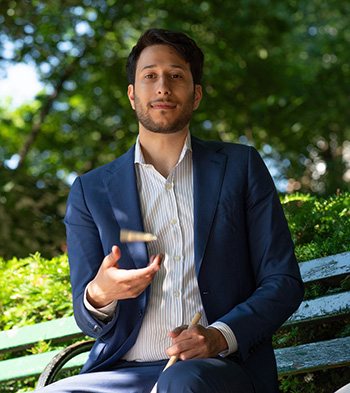 Man in blue sportcoat sits on park bench while tossing a drum stick into the air.