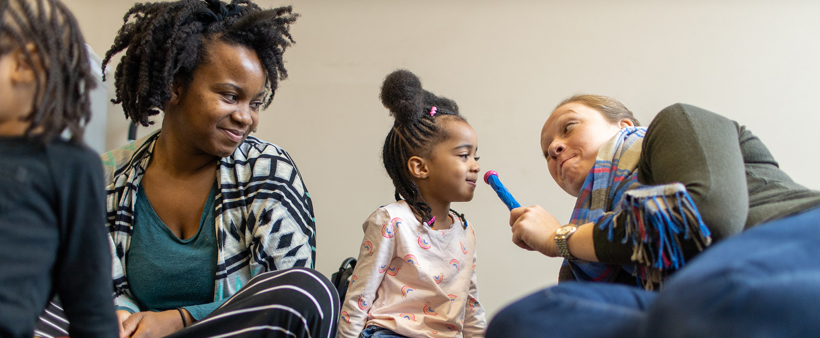 Young mom, left, watches and smiles at young child as she leans toward a play microphone held by a teacher on the right.