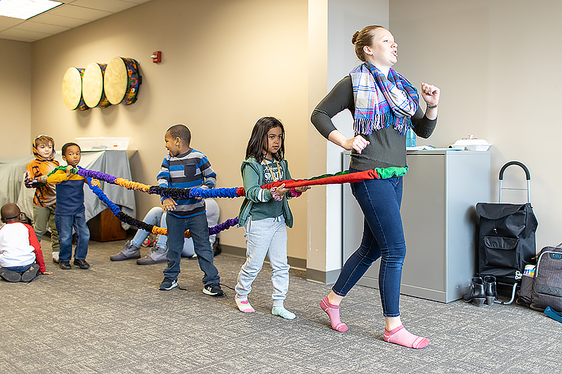 a teacher leads a march with four children following, all with one long and colorful ribbon wrapped around their waists keeping them connected. The room has three large drums on the wall behind the children.