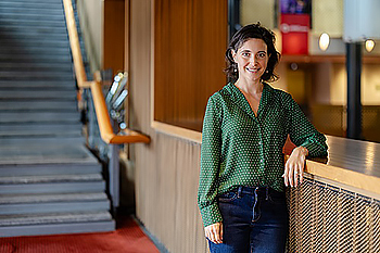 woman with short dark hair and green sweater leans her left elbow on a railing with in a theater with a staircase in the background.