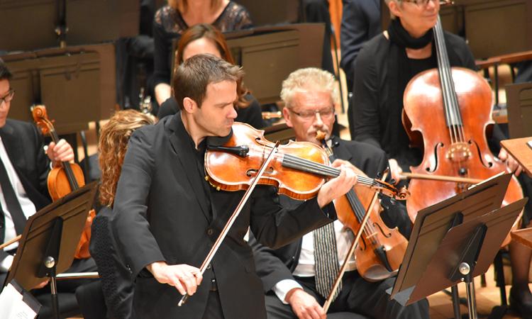 an orchestra performs while a man playing viola stands for a solo.