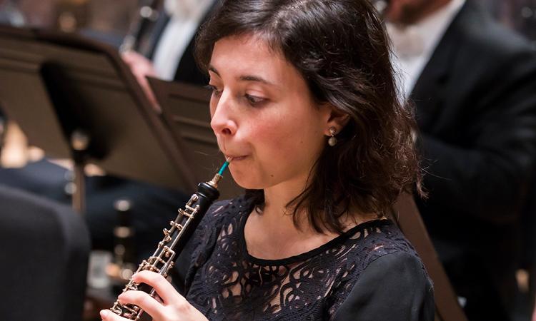 close up; woman with short dark hair performs on oboe; other musicians in background.