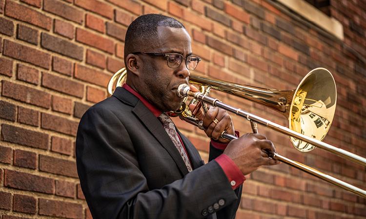 man in dark suit and glasses plays trombone while in front of a red brick wall.