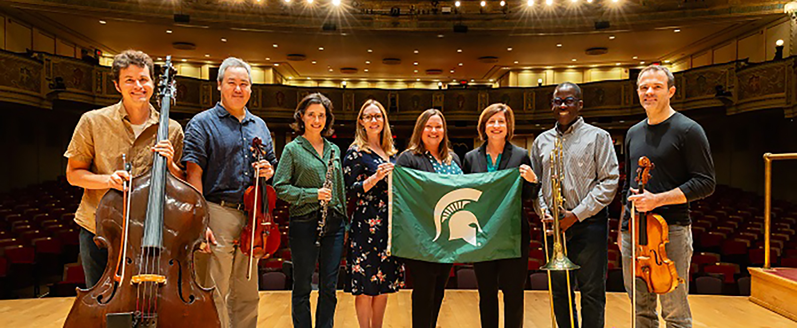 Eight musicians stand on stage, side by side with their backs to empty theater seats. From left, a man holds an upright bass, second from left a man holds a viola. Third from left a woman holds an oboe. Fourth from left, center, and third from right, three women hold a green flag with the white Michigan State University helmet emblem on it. Second from right a man holds a trombone. Far right, a man holds a viola. All people are smiling.