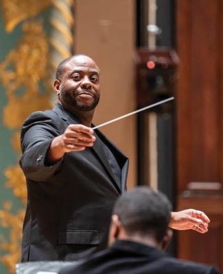 Conductor, on stage, looks into camera as he waves the baton.