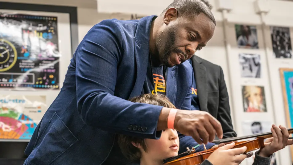 In a classroom, a teacher standing behind a young student with a violin points to the instrument while explaining something.