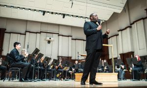 conductor stands on stage with his back to an orchestra and speaks on a microphone.