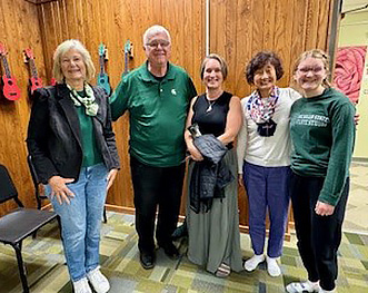 five people, four women and one man who is second from left, stand side by side in a room with green carpeting and light brown paneled wall. 