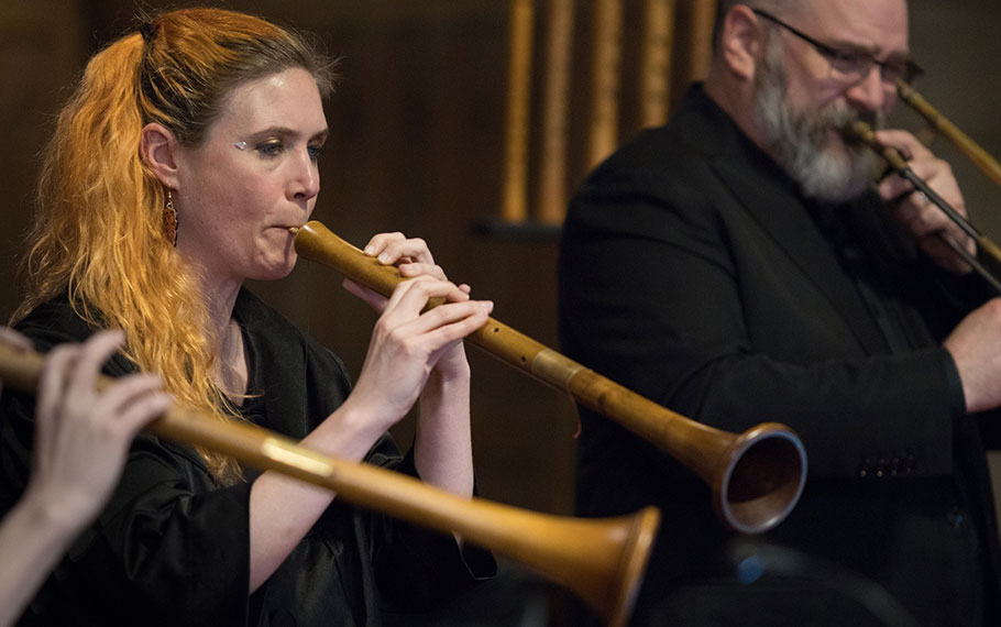 Musicians playing traditional Renaissance wind instruments during a performance, with focus on a woman playing a wooden instrument in the foreground.