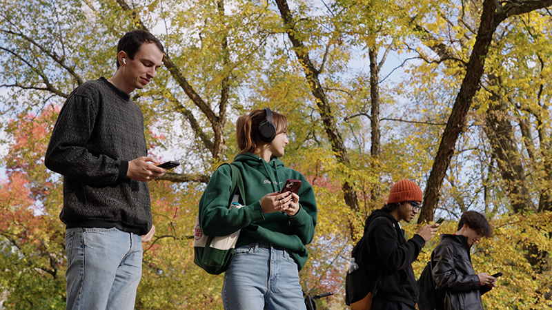 four students stand outdoors in a wooded area, three looking at their phones, one looking off into the distance, all four with earbuds or headphones.