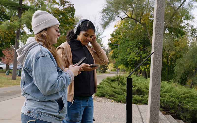 two young women stand outdoors on a sidewalk, bushes and trees in the background, as they listed to something on earbuds.