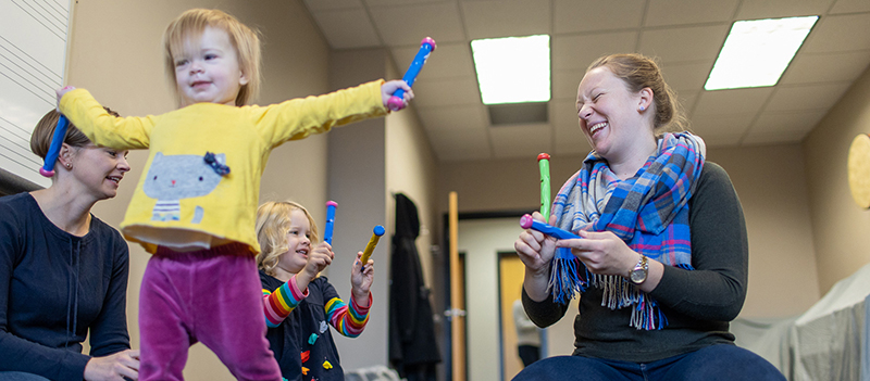 Young child smiles with outstretched arms while adult woman smiles and plays along. Another woman and small child are doing the same in the background.